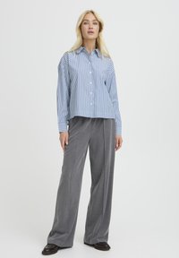 Frau steht vor einem weißen Hintergrund und trägt ein blau-weiß gestreiftes Hemd, graue weit geschnittene Hose und schwarze Schuhe.