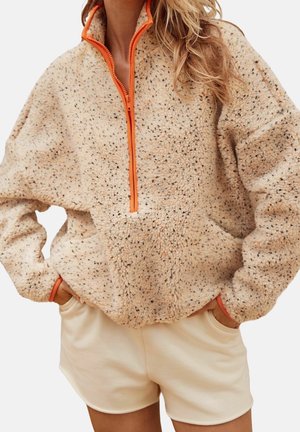Donna che indossa un pullover in pile beige maculato con cerniera e polsini arancioni, abbinato a pantaloncini color crema, mani nelle tasche.