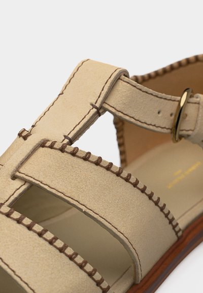 Κοντινό πλάνο της μπεζ λουρίδας σανδαλιού με καφέ ραφή και χρυσή αγκράφα σε λεία δερμάτινη σόλα.