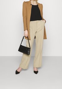 Kobieta w brązowej marynarce, czarnej bluzce, beżowych spodniaczkach w prążki, czarnych, wysmuklających butach, trzymająca małą, czarną, pikowaną torebkę w lewej ręce.