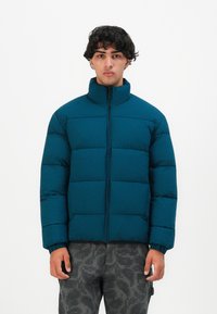 Joven de pie con una chaqueta azul acolchada con cremallera frontal y pantalones de camuflaje gris contra un fondo blanco liso.