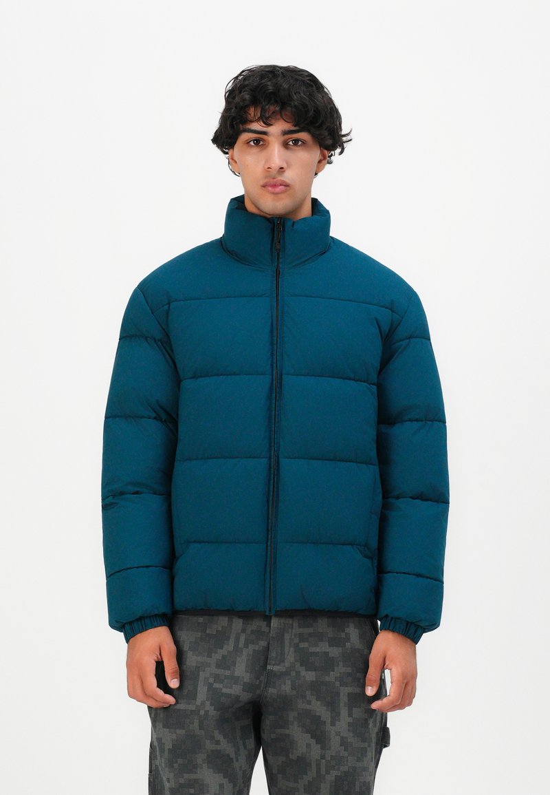 Joven de pie con una chaqueta azul acolchada con cremallera frontal y pantalones de camuflaje gris contra un fondo blanco liso.