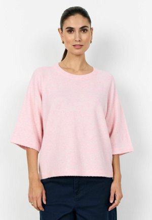 Tumejuustega naine, kandes heleroosat lahtist kampsunit küünarvarte pikkuste varrukatega ja tumedaid pükse, vaatega otse edasi.