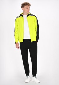 Jachetă ușoară galben-neon, cu accente negre, fermoar și glugă, purtată cu pantaloni de trening negri și adidași albi.