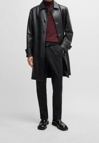 Zwarte leren jas met een knoopsluiting, gedragen over een bordeauxkleurige gestructureerde trui, gecombineerd met op maat gemaakte zwarte broek en zwarte schoenen.