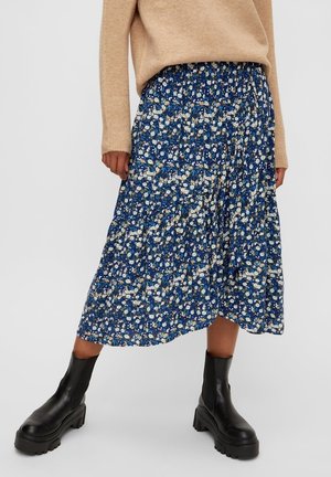 Vrouw die een beige trui, een blauwe midi-rok met bloemenprint en zwarte chunky enkellaarzen draagt, staand tegen een effen achtergrond.