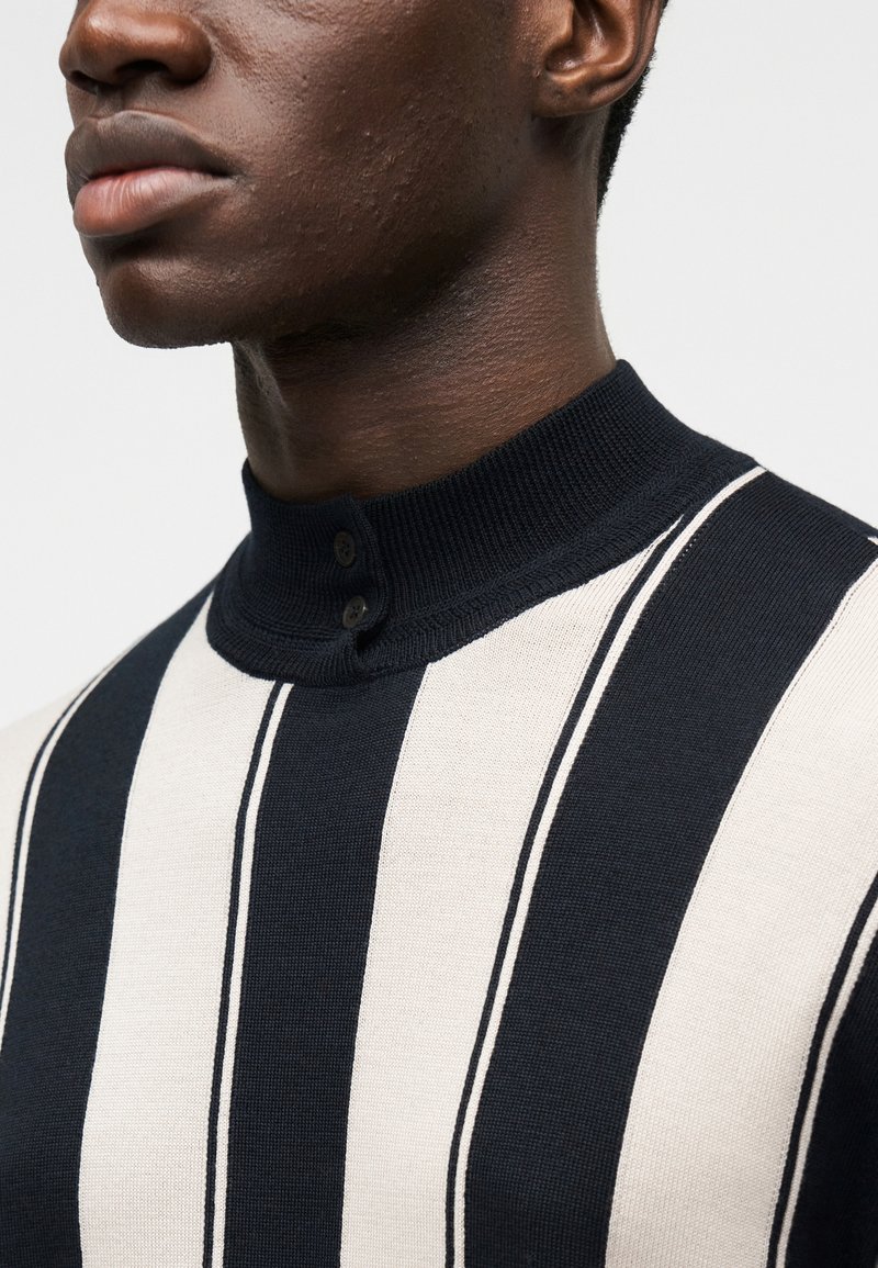 Polo en maille avec un col montant, présentant des rayures verticales alternées bleu marine et crème, et deux boutons marron sur le col.