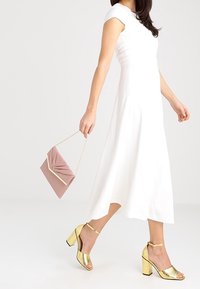 Weißes ärmelloses Kleid mit A-Linie-Silhouette; metallisch goldene Blockabsatzsandalen; rosa Clutch mit gefälteltem Design und goldenen Akzenten.