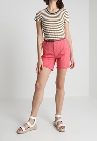 Camisa de manga corta a rayas en beige y marrón, combinada con pantalones cortos rosas. El modelo lleva sandalias blancas de tiras con suela de yute.