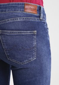 Blå denimjeans med en brun läderpatch på den bakre midjan. Har en bakficka med en silvernitat och subtila texturdetaljer.