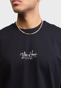 Mand iført en sort T-shirt med hvid og beige tekst "Yee Haw Unbranded Outlaw Redefined Rebel" og en guldkæde.