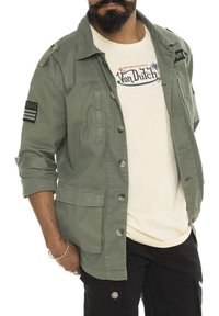 Veste en coton vert olive avec des boutons, poches sur la poitrine et un patch de drapeau américain sur la manche, portée par-dessus un t-shirt beige Von Dutch.