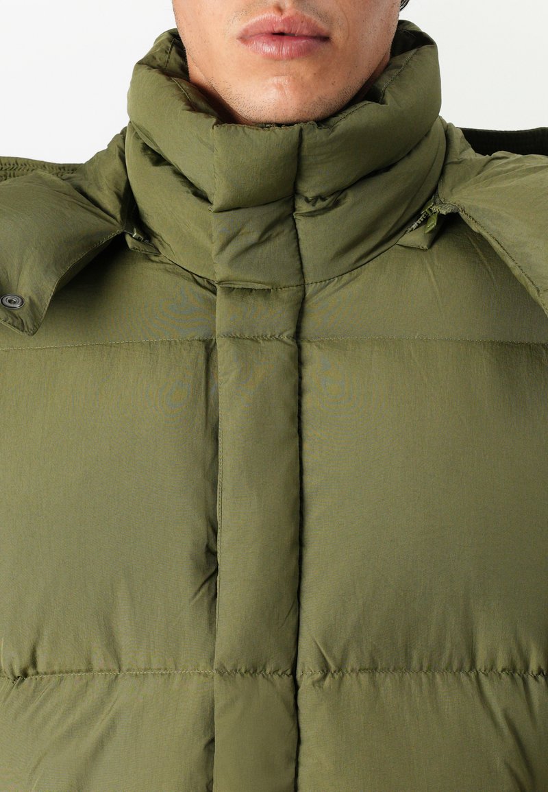 Hombre vestido con una chaqueta acolchada de color verde oliva, con un cuello alto y un cierre frontal oculto con cremallera y botones.