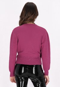 Maglione a maglia fucsia con orlo a costine e maniche lunghe, decorato con paillettes sparse per un effetto texture. Abbinato a pantaloni neri aderenti e lucidi.