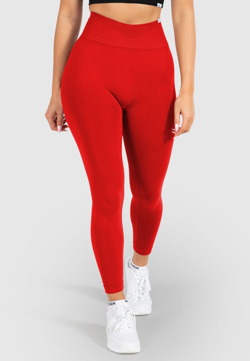 Leggings rossi a vita alta realizzati in tessuto elasticizzato, con una texture liscia e una forma aderente, abbinati a scarpe da ginnastica bianche.