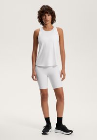 Top atletico bianco senza maniche con scollo rotondo abbinato a pantaloni bike bianchi. Scarpe da ginnastica nere con calzini neri completano l'outfit.