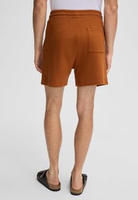 Bruine shorts van textielmateriaal, met een elastische tailleband en een enkele achterzak. Draagt samen met zwarte/slipper sandalen.