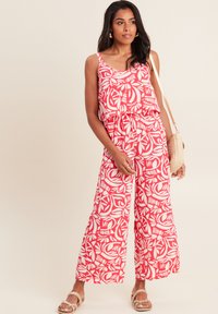 Une combinaison à motifs rouges et blancs avec un design fluide, des jambes larges et un haut à volants. Le mannequin tient un sac en osier et porte des sandales à lanières.