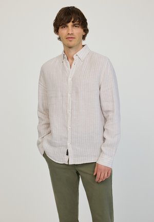 Nuori mies, jolla on ruskeat hiukset, pukeutuneena vaalean beigen raidalliseen nappipaitaan ja oliivinvihreisiin housuihin, seisoo yksinkertaisen taustan edessä.