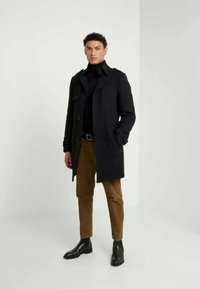 Abrigo azul marino oscuro con un diseño estructurado, detalles de botones y bolsillos laterales, combinado con un suéter negro de cuello alto y pantalones color tostado. Botas de cuero negras.