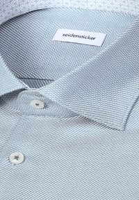 Chemise habillée bleu clair texturée avec col pointu, boutons blancs et étiquette de marque "seidensticker" à l’intérieur du col.