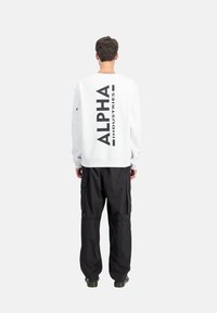 Bela gaura s kapuco z napisom "ALPHA INDUSTRIES" v črni barvi natisnjenim navpično na hrbtni strani. V kombinaciji z ohlapnimi črnimi cargo hlačami.
