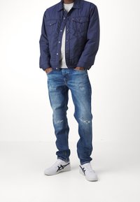 Marineblå quiltet jakke over en grå sweater, kombineret med lyseblå slidte jeans og hvide sneakers med sorte detaljer.