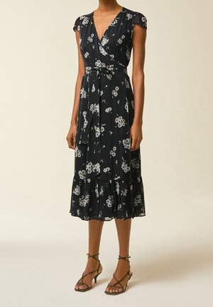 Nainen pukeutuneena mustaan midi-mittaisen kietaisumekkoon, jossa on valkoinen kukkakuosi, lyhyet hihat, vyötärölle solmittava nauha ja remmillä varustetut korolliset sandaalit.