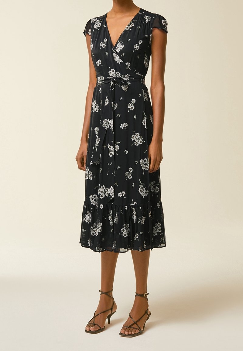 Femme portant une robe portefeuille midi noire à imprimé floral blanc, manches courtes, ceinture à nouer, et sandales à talons avec lanières.