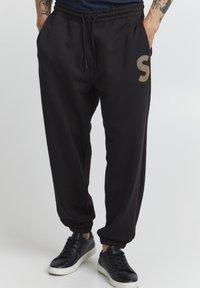Zwarte sweatpants met een elastische tailleband, zijzakken en geribde boorden; heeft een groot gouden "S"-logo op de linker dij.