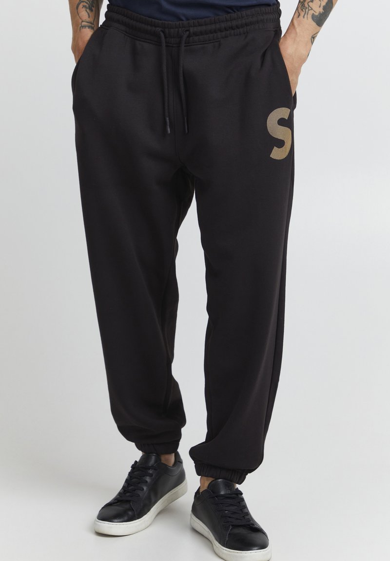 Zwarte sweatpants met een elastische tailleband, zijzakken en geribde boorden; heeft een groot gouden "S"-logo op de linker dij.