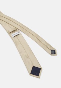 Corbata a rayas beige con una textura suave, que presenta un acento azul marino en la punta. Etiqueta de la marca adjunta cerca del extremo estrecho.