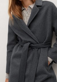 Femme portant un manteau en laine gris foncé ceinturé par-dessus un pull en maille gris clair à boutons, la main dans la poche du manteau.