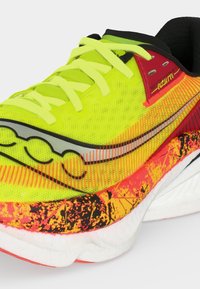 Κοντινή λήψη ενός αθλητικού παπουτσιού σε νέον κίτρινο και κόκκινο με σχεδιασμένη ενδιάμεση σόλα, κίτρινα κορδόνια και μαύρη εσωτερική επένδυση σε λευκό φόντο.