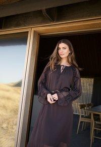 Duga, lepršava tamna haljina s naboranim rukavima i vezicom oko vrata. Tkanina izgleda lagano i blago prozirno. Remen naglašava struk.