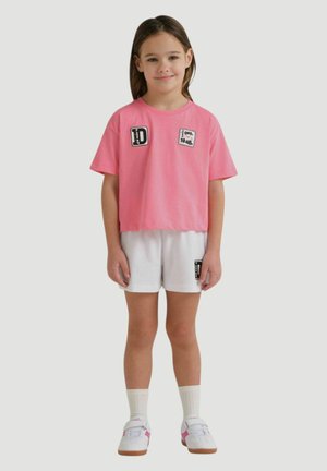 Ragazza giovane in piedi che indossa una t-shirt rosa con toppe, pantaloncini bianchi con una toppa, calzini bianchi e scarpe da ginnastica bianche con dettagli rosa.