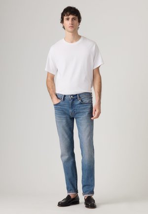 Jeune homme aux cheveux courts et foncés portant un t-shirt blanc uni, un jean bleu et des mocassins noirs, debout devant un fond neutre.
