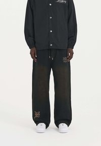 Chaqueta negra con botones a presión sobre pantalones de mezclilla oscuros y sueltos, que presentan un ligero desgastado y acentos bordados en colores contrastantes. Zapatillas blancas.