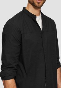 Chemise noire à manches longues avec col montant, présentant un motif texturé subtil, fermeture à boutons sur le devant et manches retroussées.