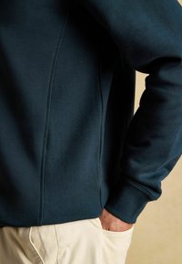 Vīrieša roka bēša biksēs, ģērbts tumši zilā garajā piedurkņu kreklā ar redzamām šuvēm un ribotām aprocēm uz rokas.