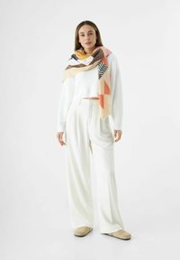 Pull en mélange de laine blanc, coupe oversize. Pantalons larges crème. Écharpe à motifs géométriques multicolores avec des nuances de rose, d'orange et des accents noirs.