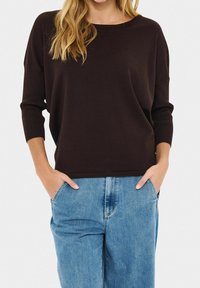 Femme aux cheveux blonds longs portant un haut à manches longues marron foncé et un jean en denim bleu clair, les mains dans les poches.