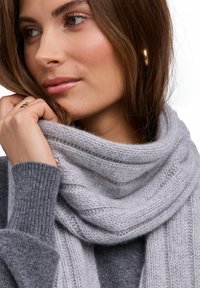 Szary dziergany szalik o miękkim, teksturowanym wykończeniu, opadający wokół linii szyi, uzupełniający szary sweter. Widoczne subtelne wzory w prążki.