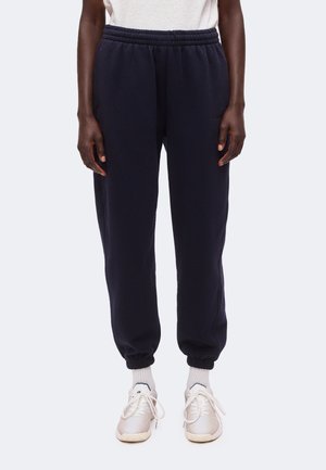 Personne portant un jogging bleu marine à taille élastique avec des chevilles élastiques, des chaussettes blanches et des baskets blanches à lacets, debout devant un fond uni.