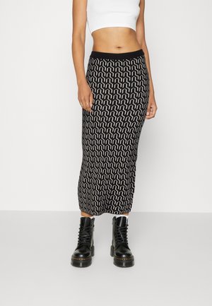 Vrouw draagt een zwart-wit geometrisch patroon midi-rok, witte cropped top en zwarte vetercombatboots, staand tegen een effen achtergrond.