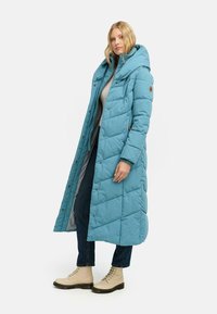 Jasnoniebieski, długi płaszcz puchowy z kapturem i wzorem w diamenty. Posiada zapięcia na guziki oraz luźny krój, zestawiony z beżowymi butami na sznurówki.