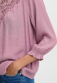 Lyserød bluse med en blomsterblonde ved halsen, afslappet pasform og flæser ved ærmerne; lavet af et blødt, struktureret stof.