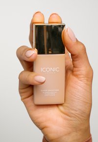 El icónico tinte para la piel blurring Super Smoother de London en un frasco de vidrio esmerilado con una tapa dorada brillante, sostenido en una mano con esmalte de uñas en un tono neutro.