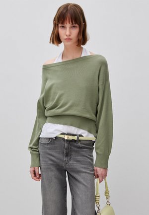 Junge Frau mit kurzen braunen Haaren trägt einen olivgrünen Pullover mit schulterfreiem Schnitt, ein weißes Oberteil, graue Jeans und hält eine blassgelbe Handtasche.