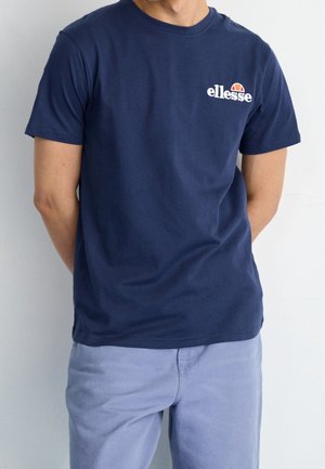 Mand iført navy blå ellesse t-shirt og lyseblå bukser, står med hænderne bag ryggen mod en ensfarvet baggrund.